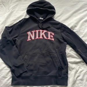 Vintage Svart Nike hoodie - Svart hoodie från Nike med stor broderad NIKE-logga i grått och rött på bröstet. Klassisk känguruficka framtill, huva med snörning och ribbade muddar. Perfekt för chill eller streetwear. Swoosh-detalj på ärmen.