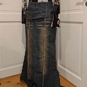 Y2k maxi jeanskjol med bälte  - Unik lång jeanskjol i blå denim med slitna detaljer, råa kanter och markerade sömmar. Kjolen har ett brett denim-bälte med stor spänne och snörning längs sidorna. Dragkedja bak och coola slitningar framtill.