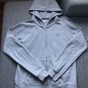 Ljusgrå hoodie från GANT med dragkedja - Snygg ljusgrå hoodie från GANT med huva och hel dragkedja framtill. Tröjan har två fickor, ribbade muddar och ett litet broderat GANT-logo på bröstet. Perfekt för chill dagar och enkel att matcha med jeans eller joggers.