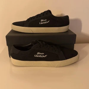 Svarta Saint Laurent sneakers - Svarta sneakers från Saint Laurent med vit broderad logga på sidan. Skorna har en klassisk låg profil, snörning och vit platt gummisula. Ovandelen är i textil och mocka för en stilren och clean look. Perfekta för dig som gillar minimalistisk design.
