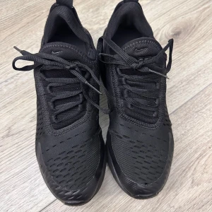 Nike Air Max 270 helsvarta sneakers - Nike Air Max 270 sneakers i helsvart färg med mesh-överdel och synlig Air-enhet i hälen. Snygg sportig design med snörning och diskreta Nike-loggor på sidan och tungan. Skorna har rund tå och platt sula, perfekt för dig som gillar streetstyle och komfort.