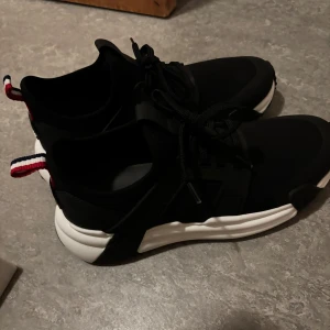 Svarta sneakers från Moncler - Snygga svarta sneakers från Moncler med vit chunky sula och klassisk snörning. Skorna har en sportig design med Moncler-logga på innersulan och hälen samt en randig flik i rött, vitt och blått baktill. Ovandelen är i textil och syntet för en modern look. Köpt för 5400kr kvitto finns och moncler påse kommer med samt box (äkta)