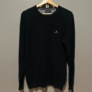 Svart stickad tröja från GANT - Svart stickad tröja från GANT i 100% bomull. Klassisk rund hals och diskret logga broderad på bröstet. Långärmad modell med ribbade muddar vid ärmslut och nederkant. Perfekt för dig som gillar stilrena och enkla plagg.