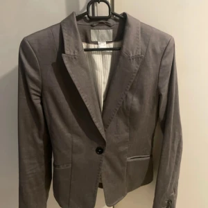 Grå kavaj från H&M med knappar - Snygg grå kavaj från H&M med klassisk krage, tre dekorativa knappar vid ärmslut och en knapp framtill. Fodret är ljusgrått med tunna ränder. Figursydd modell med fickor och detalj baktill. Perfekt för en stilren look.