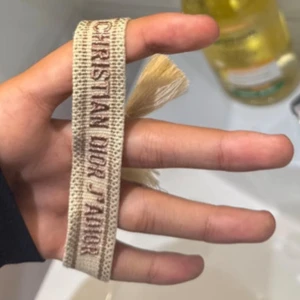 Christian Dior J'Adior armband beige - Snyggt beige armband från Christian Dior med broderad text 'J'Adior' i brunt. Armbandet har justerbar knytning och dekorativa tofsar i ändarna. Perfekt accessoar för att lyfta din outfit med en lyxig touch.
