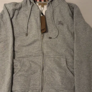 Grå hoodie från Burberry med dragkedja - Snygg grå hoodie från Burberry med klassiskt rutigt foder i huvan och insidan. Tröjan har dragkedja, snörning i huvan och broderad logga på bröstet. Perfekt för dig som vill ha en stilren och lyxig look med streetkänsla. Säljer då den ej passar helt ny oanvänd det går o lämna tillbaka om den ej ser ny ut. Storlek L men kan tänka mig M 