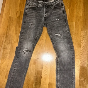 Grå slitna jeans från Jack & Jones - Säljer ett par grå jeans från Jack & Jones med slitningar och trasiga detaljer på benen. Jeansen har fem fickor, smal passform och klassisk knappgylf. Perfekta för en avslappnad och trendig look.