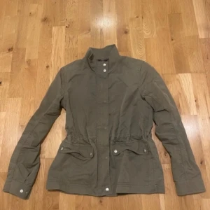 Olivgrön vindjacka från GANT - Snygg olivgrön vindjacka från GANT med hög krage, tryckknappar och två stora fickor framtill. Jackan har dragsko i midjan för en schysst passform och är tillverkad i bomull. Perfekt för vår och höst när du vill ha en stilren look. Str , 164 passar xs- s 