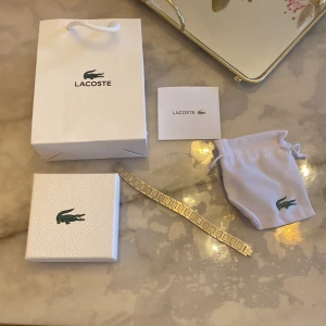Lacoste armbandsur i silver - Snyggt armbandsur från Lacoste med länkarmband i silverfärgat metall. Länkarna har graverade detaljer och Lacostes ikoniska krokodillogga. Klockan levereras med originalförpackning, påse och tygpåse med logga.