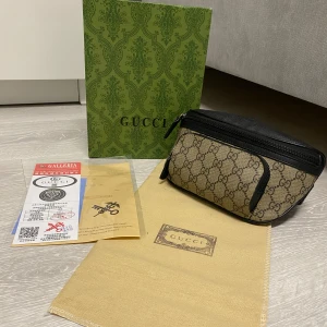 Gucci väska  - Säljer en snygg beige Gucci väska med klassiskt GG-mönster och svarta skinn-detaljer. Väskan har dragkedja upptill och ett extra fack framtill. Perfekt för dig som vill ha en stilren och lyxig accessoar.