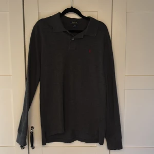 Mörkgrå pikétröja från Polo Ralph Lauren - Säljer en mörkgrå långärmad pikétröja från Polo Ralph Lauren i storlek XL i barn storlek . Tröjan har klassisk krage, knäppning vid halsen och den ikoniska röda loggan broderad på bröstet. Perfekt för dig som gillar stilren och tidlös design. Nypris 2000kr