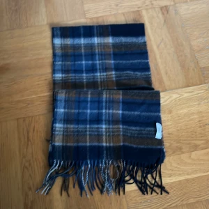 Rutig blå halsduk från Bläck - Snygg halsduk från Bläck i rutigt mönster med marinblå, grå och bruna toner. Mjuk och varm känsla, perfekt att slänga runt halsen när det är kallt ute. Fransar i ändarna ger en chill vibe och den passar till de flesta jackor.