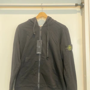 Svart hoodie från Stone Island - Svart hoodie från Stone Island med klassisk logotyp-patch på vänster ärm. Tröjan har huva med snörning, hel dragkedja framtill och rymliga fickor. Tillverkad i mjuk bomull för skön komfort. Perfekt för dig som gillar streetwear och snygga detaljer.