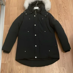 Svart dunjacka Moose Knuckles med pälskrage - Svart dunjacka från Moose Knuckles med snygg silvrig logga på ärmen och vit pälskantad huva. Jackan har silverfärgade knappar, dragkedja och längre bakstycke. Perfekt för kalla dagar och har en cool broderad detalj på insidan.