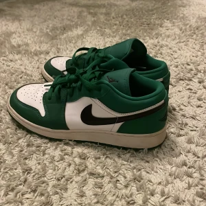 Nike Air Jordan 1 Low grön/vit - Säljer ett par Nike Air Jordan 1 Low i grön och vit färg med svarta detaljer. Skorna har klassisk snörning, rund tå och den ikoniska swoosh-loggan på sidan. Tillverkade i skinn med perforerad tåbox för extra andningsförmåga. Perfekta för dig som vill sticka ut med din sneakerstil.