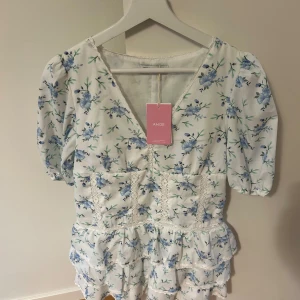 Blommig kortklänning från Design by Amor - Supersöt vit kortklänning från Design by Amor med blått blommönster, v-ringning och puffärmar. Klänningen har volangdetaljer och spets längs sömmarna, vilket ger en romantisk vibe. Perfekt för sommarens alla tillfällen! Aldrig använd då den inte passade 