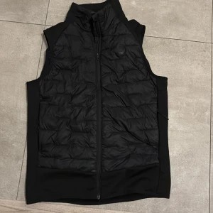 Svart dunväst från Everest strl 40 - Svart dunväst från Everest med quiltad framsida och diskret logga på bröstet. Västen har hög krage och dragkedja framtill. Materialet är syntet och dun, perfekt för lager-på-lager under kyliga dagar. Snygg och sportig look som funkar till många stilar.