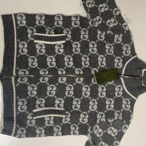 Grå kofta med GG-mönster från Gucci - Grå stickad kofta från Gucci med vitt GG-mönster över hela plagget. Koftan har dragkedja framtill, ribbade muddar och två fickor med vit kant. Hög krage och exklusiv känsla, perfekt för dig som gillar lyxiga detaljer.