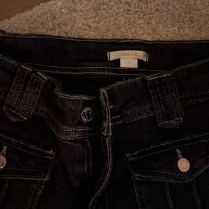 Svarta jeansbyxor från Gina Tricot - Svarta jeansbyxor från Gina Tricot med raka ben och coola fickor framtill med lock och silverfärgade knappar. Byxorna har breda hällor och klassisk dragkedja. Perfekta för dig som gillar en enkel men snygg stil.