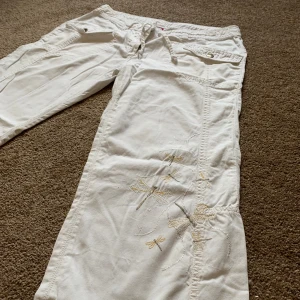 Vita cargopants från H.H.S med broderi - Super gulliga vita cargopants från H.H.S med dragsko i midjan och vid benslut. Byxorna har snygga fickor med knappar och broderade trollsländor i ljusa toner på ena benet. Tillverkade i bomull för en skön och avslappnad känsla. Perfekta för en chill och trendig look.