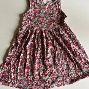 Blommig ärmlös klänning från H&M - Supersöt ärmlös klänning från H&M med blommigt mönster i rött, rosa och grönt på vit botten. Klänningen har en rund halsringning och ett utställt kjolparti som ger en skön och ledig känsla. Perfekt för varma dagar och somriga vibes!