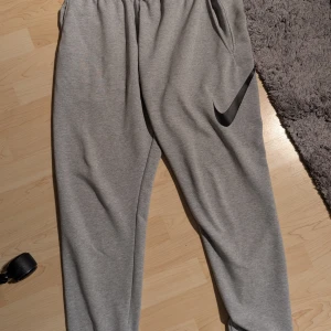 Grå mjukisbyxor från Nike - Säljer ett par grå mjukisbyxor från Nike med svart logga på benet. Byxorna har elastisk midja med snörning och sidofickor. Perfekta för chill eller träning, riktigt sköna och stilrena.