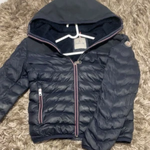 Mörkblå Moncler jacka med huva - Snygg mörkblå jacka från Moncler med broderad logga på ryggen och patch på ärmen. Jackan har huva, dragkedja framtill och quiltad design. Fodrad insida för extra värme och coola detaljer med rödvit rand vid dragkedjan. Passar M också
