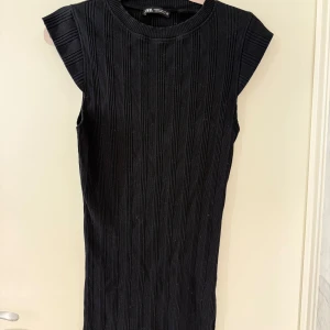 Svart ribbad kort klänning från Zara - Svart ribbad klänning från Zara med coola breda axlar och ärmlös design. Kort modell med rund hals och vertikala ribbade detaljer som ger en snygg struktur. 