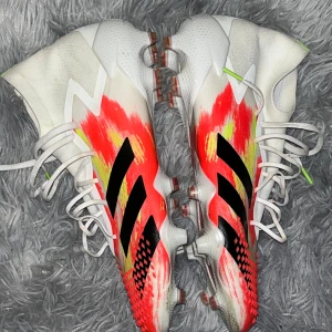 Adidas Predator 20.1 fotbollsskor - Säljer ett par Adidas Predator 20.1 fotbollsskor i storlek 44 2/3. Skorna har en cool design med vit bas och röda, gula och svarta detaljer. De har snörning, högre skaft och mönstrad ovansida för extra grepp. Perfekta för dig som vill sticka ut på planen.