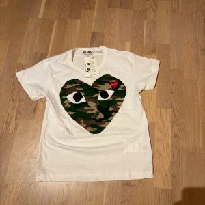 Vit Comme des Garçons Play t-shirt S - Vit t-shirt från Comme des Garçons Play med stort hjärta i camouflage på bröstet och ett litet rött hjärta med ögon. Klassisk passform och rund hals. Tillverkad i mjuk bomull, perfekt för en cool och avslappnad stil.
