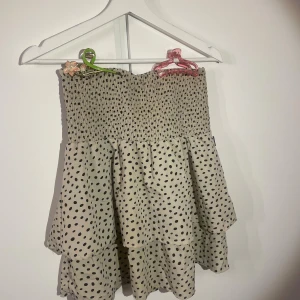 Prickig volangkjol från Seaside - Supersöt kort volangkjol från Seaside i beige med svarta prickar. Kjolen har smock i midjan för en bekväm passform och dubbla volanger som ger en flowy känsla. Perfekt för dig som gillar lekfulla och trendiga plagg.