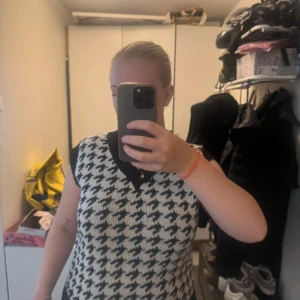 Svartvit stickad väst med hundtandsmönster - Säljer en snygg stickad väst i svart och vitt med klassiskt hundtandsmönster. Västen har v-ringning och svarta ribbade kanter vid hals, ärmhål och nederkant. Perfekt att styla över en skjorta eller t-shirt för en trendig look. Storlek S/M