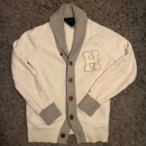 Vit kofta från Tommy Hilfiger - Vit och grå kofta från Tommy Hilfiger med stora bruna knappar och ett broderat H på bröstet. Koftan har ribbade muddar och krage i grått, samt långärmad passform. Stickad i mjukt bomullsmaterial, perfekt för chill höstkvällar.