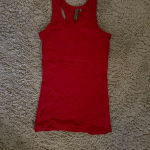 Röd linnetopp i bomull - Säljer en enkel och snygg röd linnetopp i bomull. Modellen är ärmlös med klassisk racerback och passar perfekt till sommaren. Toppen är figurnära och har en stilren look som är lätt att matcha med jeans eller shorts.