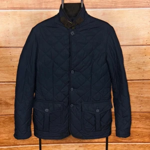 Mörkblå barbour quiltad jacka   - Quiltad mörkblå jacka från Barbour med ståkrage och snyggt spänne i halsen. Jackan har två stora fickor med knappstängning, knappar framtill och diskret Barbour-logga på insidan. Perfekt för dig som gillar klassisk stil med modern touch.