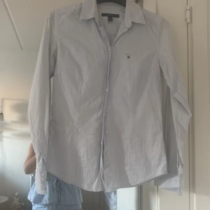 Randig skjorta från GANT i bomull - Klassisk ljusblå och vit randig skjorta från GANT med krage och knäppning framtill. Skjortan har långa ärmar med manschettknappar och liten broderad logga på bröstet. Perfekt för en clean och stilren look.