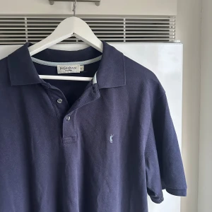 Mörkblå polo t-shirt från Yves Saint Laurent - Säljer en mörkblå polo t-shirt från Yves Saint Laurent med klassisk krage och knäppning framtill. T-shirten har broderad YSL-logga på bröstet och är tillverkad i bomull. Perfekt för dig som gillar stilrena och tidlösa plagg.