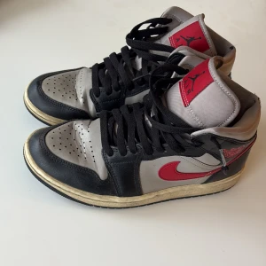 Nike Air Jordan 1 High svart/röd - Nike Air Jordan 1 High sneakers i svart, grått och rött med klassisk röd swoosh och Air Jordan-logga på sidan. Skorna har svarta snören, perforerad tå och vintage-inspirerad gul sula. Tillverkade i skinn och syntet för en ikonisk streetlook.