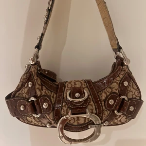 Guess vintage bag<3 - Cool axelväska från Guess med brunt GG-mönster, detaljer i blankt mörkbrunt skinn och stora silverspännen. Insidan av axelbandet är i guldigt, ormskinnsmönstrat material. Väskan har flera nitar och ett stort metallspänne framtill. Perfekt för dig som gillar statement-accessoarer. Kom gärna privat för frågor eller pris förslag!!☺️(lite små skavanker på sidorna av remmen, men inget som syns)