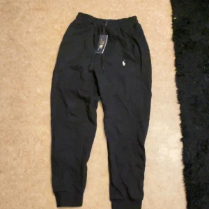 Svarta mjukisbyxor från Polo Ralph Lauren - Svarta mjukisbyxor från Polo Ralph Lauren med dragsko i midjan och klassisk logga broderad på benet. Byxorna har ribbade muddar vid bensluten och är tillverkade i mjukt bomullsmaterial för en chill och bekväm stil.