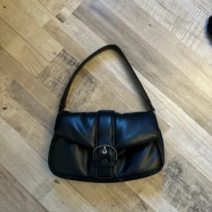 Svart axelväska med spänne - Snygg svart axelväska i skinn med stilren design och ett stort spänne framtill. Perfekt storlek för det viktigaste och enkel att slänga över axeln. Väskan har en mjuk form och klassisk look som funkar till många outfits.