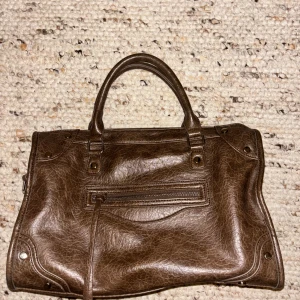 Brun handväska från Gina Tricot - Snygg brun handväska från Gina Tricot i skinnimitation med två handtag och dekorativa nitar. Väskan har en dragkedja framtill och rymligt innerfack med extra fickor. Perfekt storlek för att få plats med allt du behöver och passar till många olika stilar.