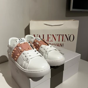 Valentino skor - Säljer ett par Valentino sneakers i vitt skinn med ljusrosa band och ikoniska nitar. Skorna har rund tå, platt sula och snörning framtill. Perfekt för dig som vill ha en lyxig och edgy look med snygga detaljer från Valentino.