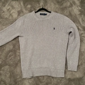 Grå srickad tröja från  Polo Ralph Lauren - Snygg ljusgrå stickad tröja från Polo Ralph Lauren med klassisk rund hals och diskret logga broderad på bröstet. Tröjan har ribbade muddar vid ärmslut och nederkant, perfekt för dig som gillar stilren och enkel design. Lite nopprig på vissa ställen, men inget som man lägger märke till. Priset är ej hugget i sten!