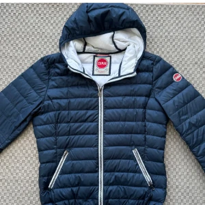 Mörkblå pufferjacka från Colmar - Snygg mörkblå pufferjacka från Colmar med huva och vita detaljer. Jackan har quiltad design, två fickor med dragkedja och Colmar-logga på ärmen. Perfekt för kyliga dagar och har en stilren look som funkar till många outfits. Bra skick, den e använd sparsamt.