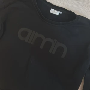 Svart croppad tröja från Aimn - Svart långärmad croppad tröja från Aimn med stor logga framtill. Mjuk och skön, perfekt för chill eller träning. Rund halsringning och stilren design som funkar till många olika looks.