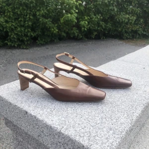 Bruna pumps med låg klack från Isabella - Eleganta bruna slingback pumps från Marbella. Bekväm klackhöjd och justerbar rem runt hälen för bästa passform. Skorna har en elegant fyrkantig tå och är tillverkade äkta skinn. Perfekta för både vardag och fest. Jättesnygga till hösten. Mycket fint skick!