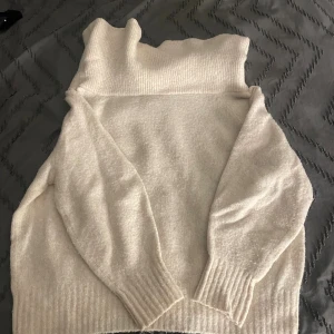 Vit stickad polotröja med bred krage - Mysig vit stickad polotröja med bred, ribbad krage som kan vikas ner. Tröjan har långa ärmar och ribbade muddar vid ärmslut och nederkant. Perfekt för kyliga dagar och enkel att styla till olika outfits.