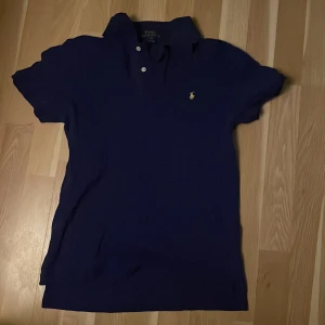 Blå pikétröja från Polo Ralph Lauren - Klassisk blå pikétröja från Polo Ralph Lauren med korta ärmar och den ikoniska broderade loggan i gult på bröstet. Tröjan har krage och knappar framtill. Perfekt för en avslappnad men ändå stilren look.