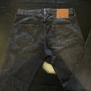 Levi's 501 svarta jeans W30 L32 - Svarta Levi's 501 jeans med klassisk rak passform och femficksdesign. De är använda 2 gånger och är i sjukt ba skick. Jeansen har knappgylf, silverfärgade nitar och den ikoniska läderpatchen bak i midjan. Tillverkade i slitstarkt bomullsjeans med en mörk tvätt som ger en cool vibe.